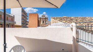 A balcony with a table and chairs, a white umbrella, and a view of buildings and a church.