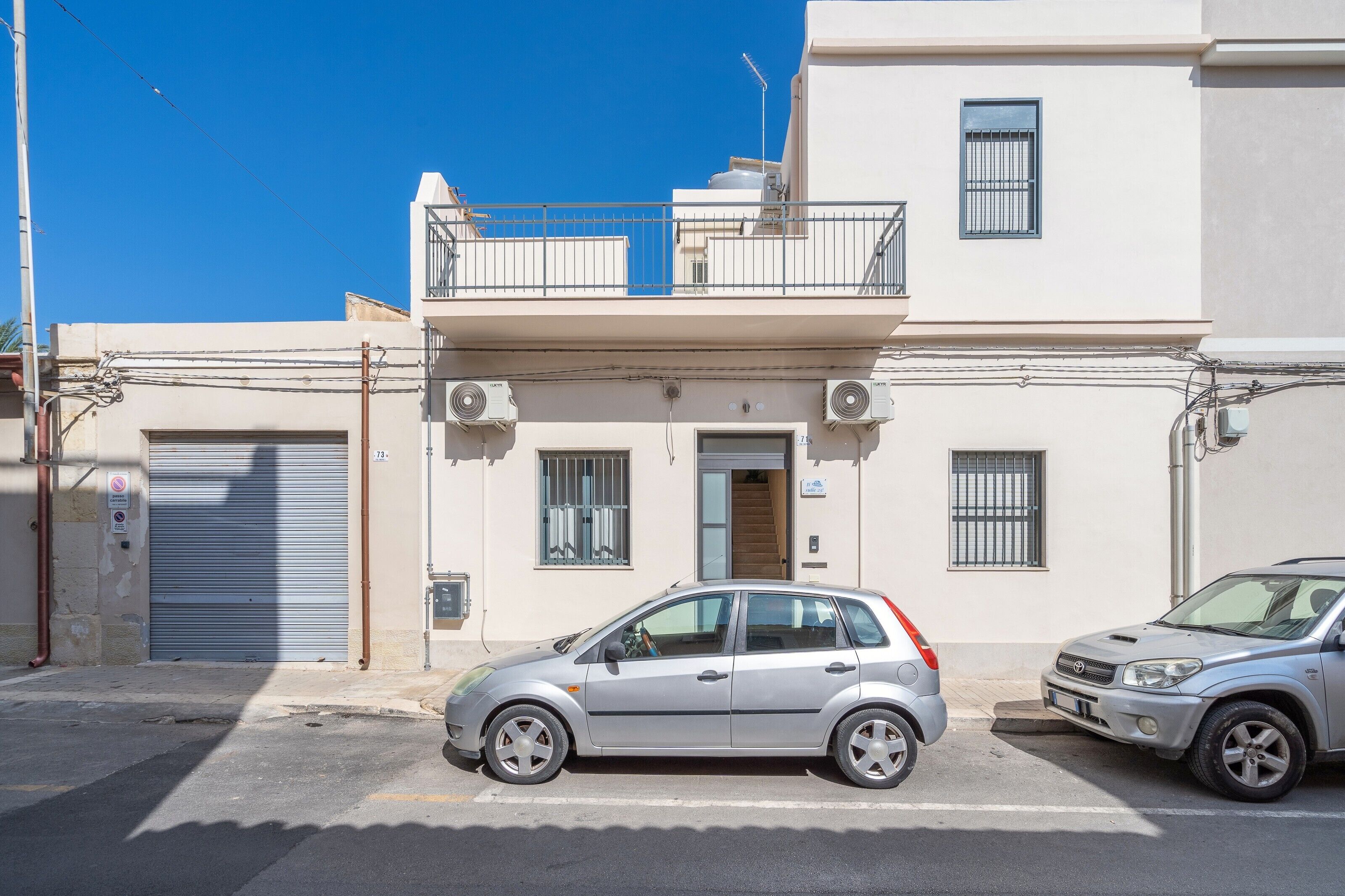 Un edificio a due piani con un balcone, un'autorimessa e due automobili parcheggiate.