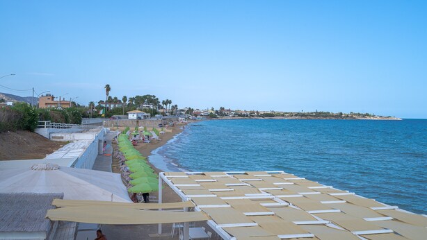 A beachside hotel with rows of sun loungers and umbrellas, a clear view of the sea, and a distant shoreline with buildings.