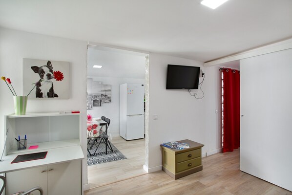A room with a desk, a television, a small cabinet, and a door leading to another room.
