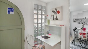 A room with a white desk, a chair, a window with a view, a wall-mounted artwork, and a door with a WiFi sign.