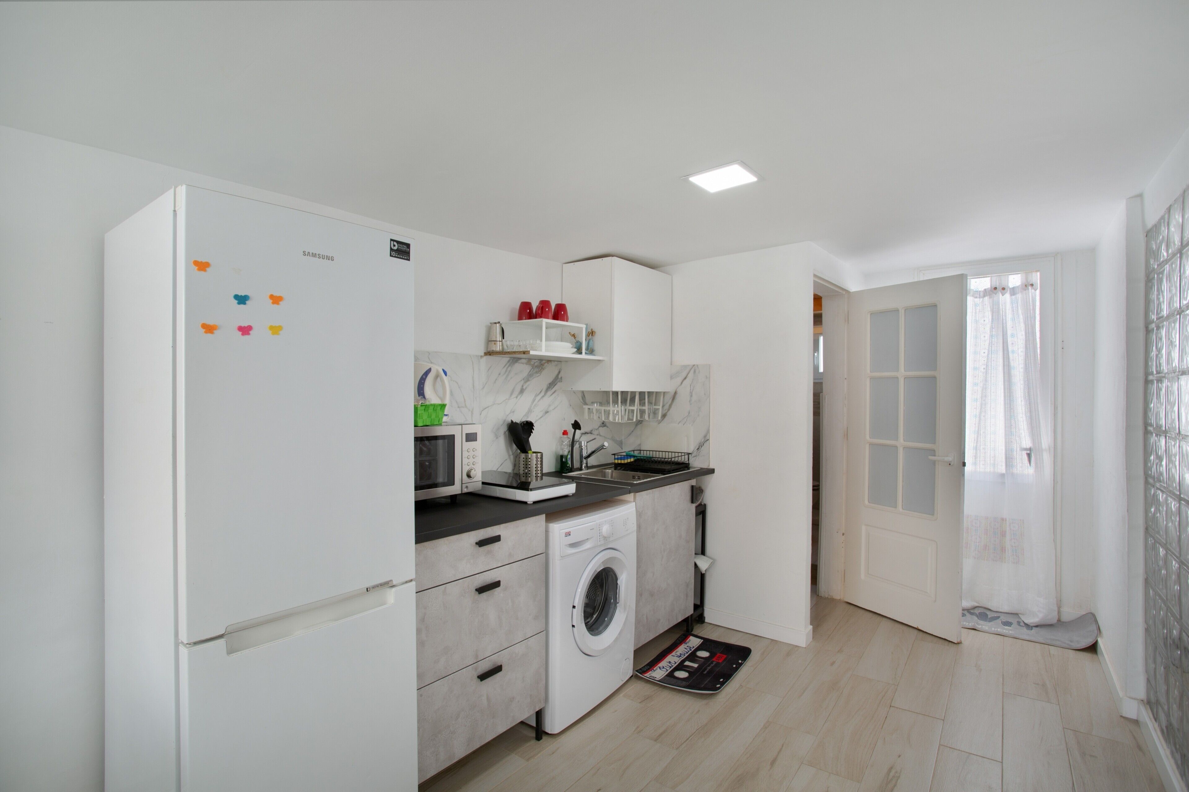 A compact kitchen with a washing machine, microwave, and a small shelf with red cups.