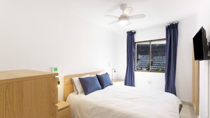 A bedroom with a bed, a wooden nightstand, a ceiling fan, a TV, and a window with curtains. - Vacation Home 'Casa Neale' with Sea View, Shared Pool and Private Terrace (Puerto Rico)