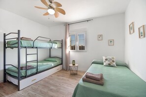 Een slaapkamer met een stapelbed, een plafondventilator, een klein nachtkastje, een raam met gordijnen en een bed met kussen.