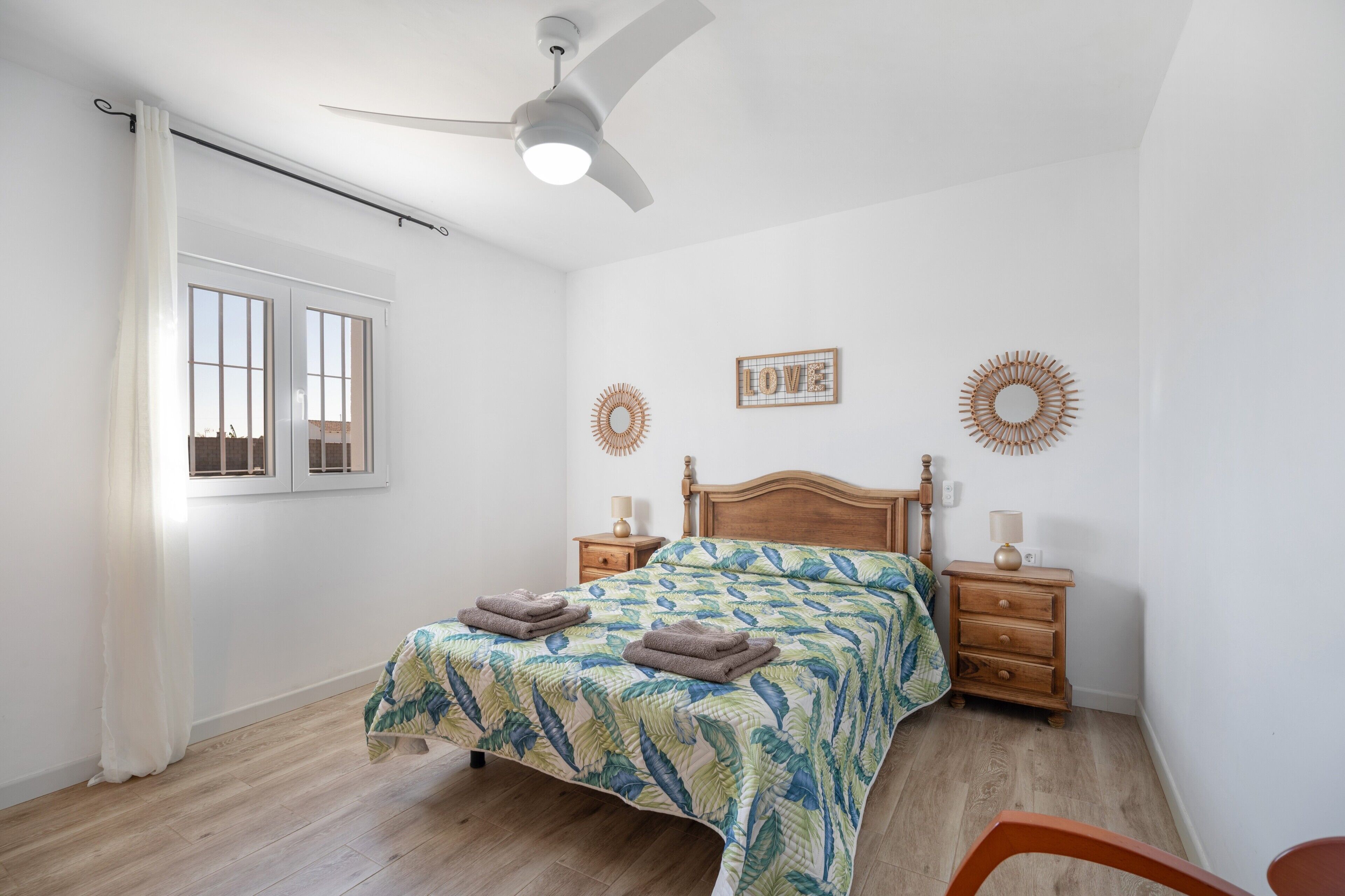 Une chambre à coucher comprenant un lit, deux tables de chevet, un ventilateur de plafond et une fenêtre avec des rideaux.