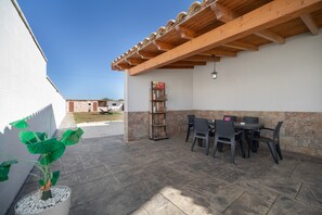 Een terras buiten met een tafel, stoelen, een bloempot en een boekenkast.
