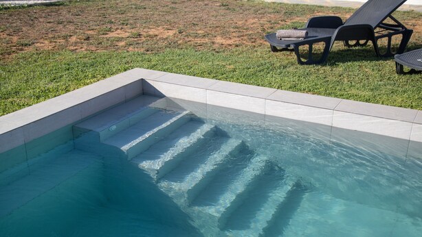 Ein Schwimmbecken mit klarem Wasser, Stufen zum hineinsteigen und zwei Liegestühle auf der Wiese.