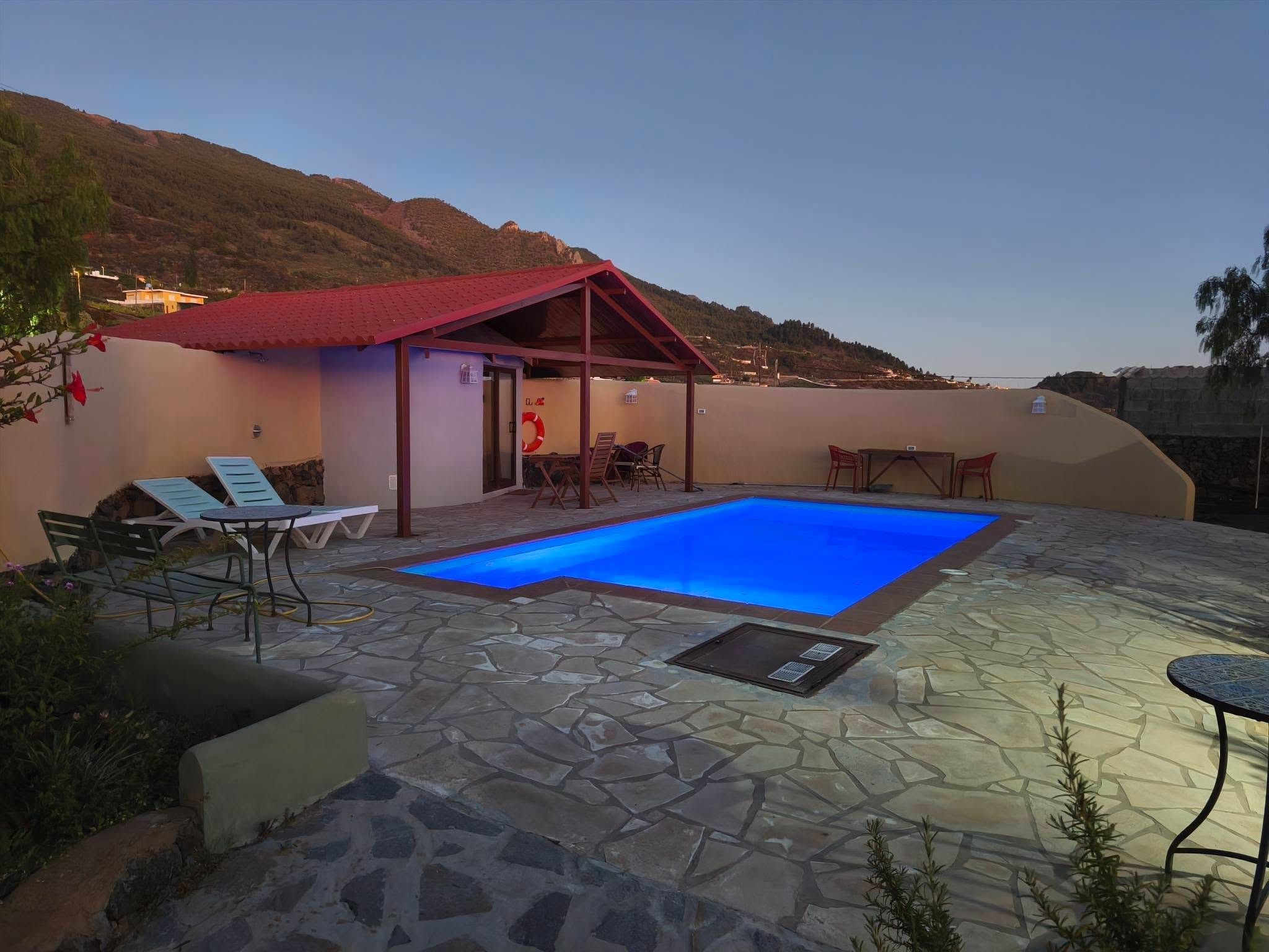 Une piscine avec une terrasse en pierre, un espace salon couvert et une vue sur les collines en arrière-plan.