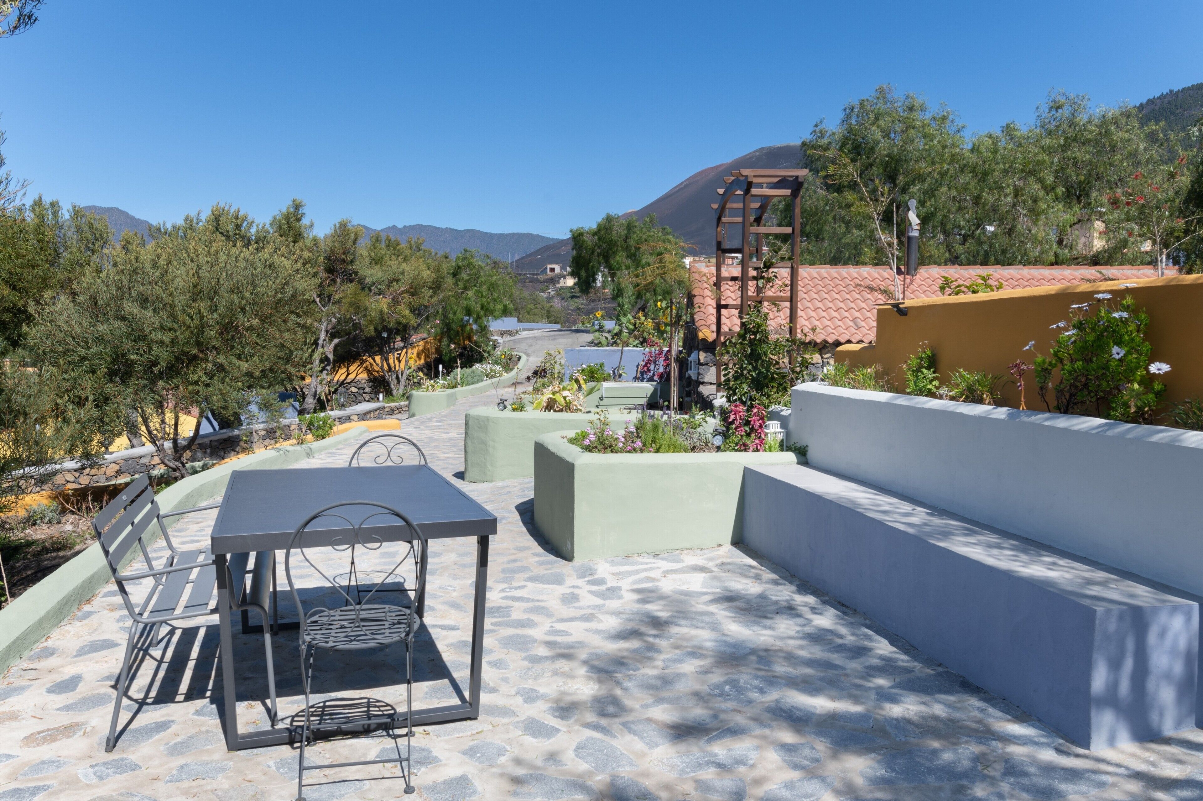 A patio with a table and chairs, surrounded by planters and a view of mountains.
