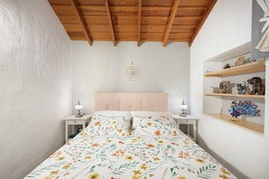 Une chambre avec un plafond en bois, un lit recouvert d’une literie à motifs floraux, des tables de chevet avec des lampes et des étagères ornées d’objets décoratifs.