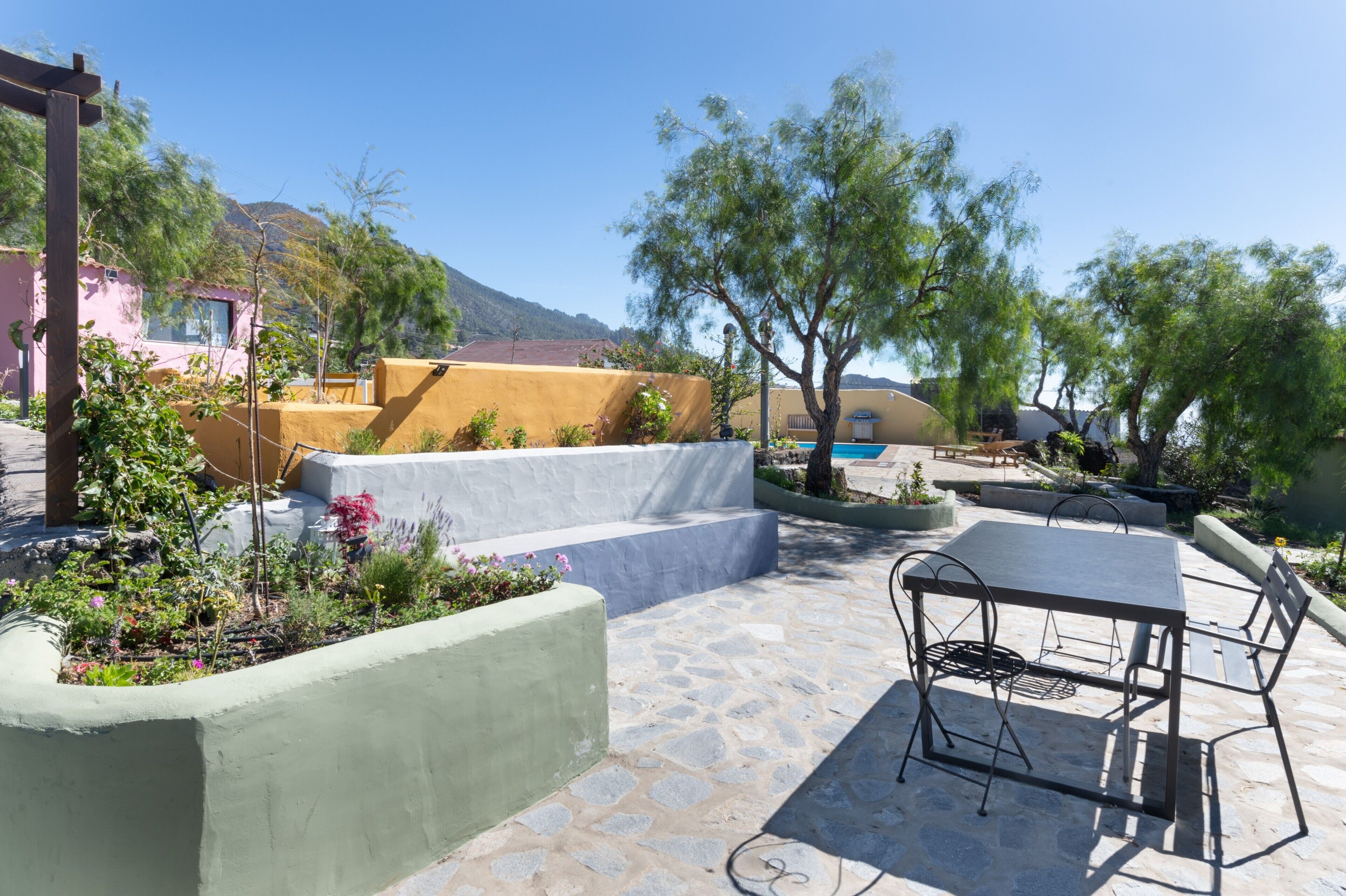 A patio with a table and chairs, surrounded by plants and a mountain view.
