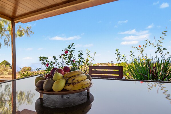 A table with a fruit bowl containing apples, pears, bananas, and a red fruit, possibly a peach, under a clear sky with some clouds and greenery in the background.