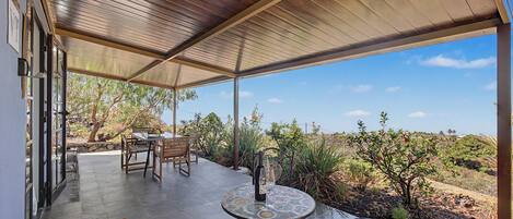 An outdoor patio with a covered seating area, a table with a bottle and glasses, and a view of the surrounding landscape.