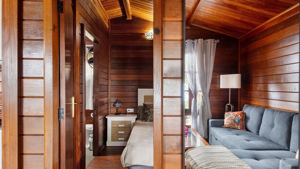 A wooden cabin interior with a bed, a sofa, a lamp, and a window with curtains.