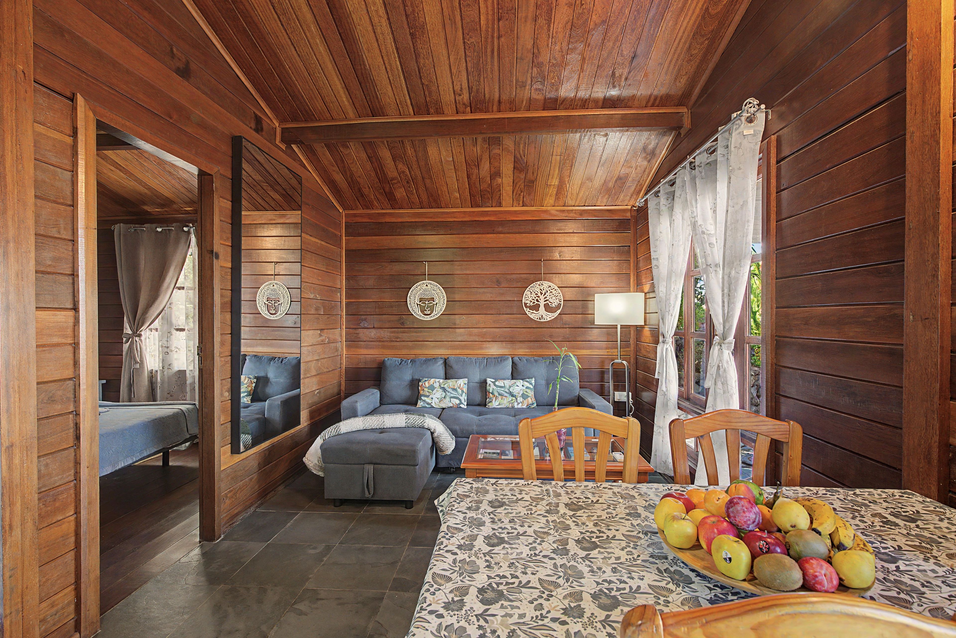 Ein gemütliches Blockhauss-Interieur mit Holzbalkendecke, einer Sofaecke, einem Esstisch mit Obst und einem Spiegel, der den Raum reflektiert.