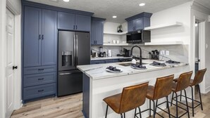 A modern kitchen with blue cabinets, a stainless steel refrigerator, a microwave, and a large island with bar stools.