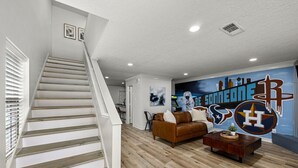 A modern living room with a staircase, a brown leather sofa, a wooden coffee table, and a mural with sports team logos.
