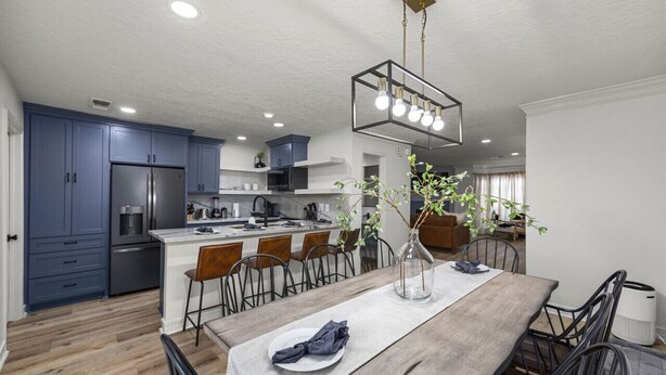 A modern kitchen with blue cabinets, a stainless steel refrigerator, and a dining area with a wooden table and chairs.