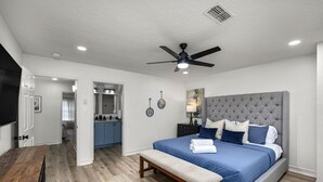 A bedroom with a large bed, a bench, a ceiling fan, and a view into the kitchen.