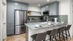 A modern kitchen with grey cabinets, stainless steel appliances, a marble countertop, and bar stools.