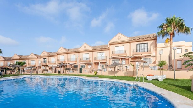 A swimming pool with a clear blue water surface, surrounded by a row of buildings with balconies and a palm tree.