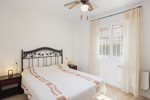 Un dormitorio con cama, mesitas de noche, ventilador de techo y ventana con cortinas.