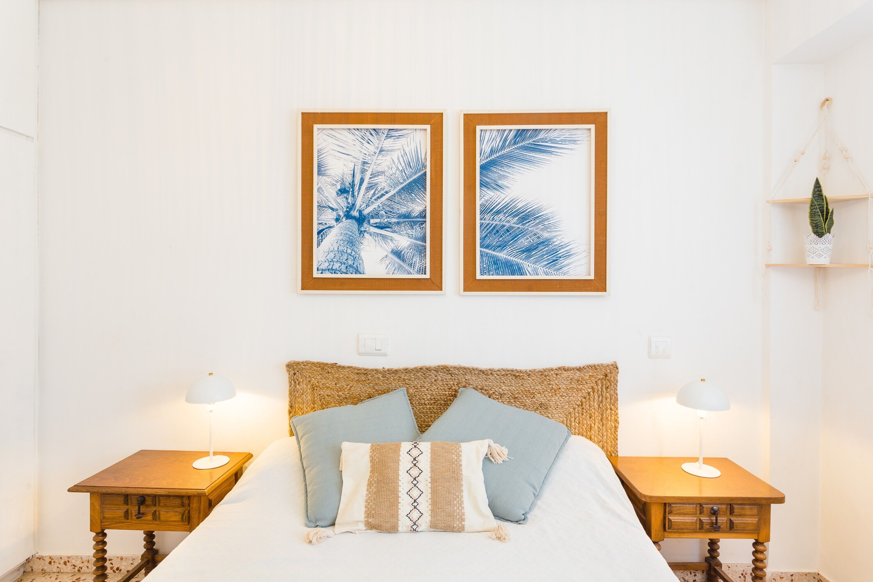 Un lit avec une tête de lit en osier, deux tableaux encadrés accrochés au mur, deux tables de chevet avec des lampes et une plante suspendue.