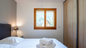 Habitación de hotel con cama, ventana que ofrece vistas a un edificio y árboles, mesita de noche con lámpara y un armario de madera.