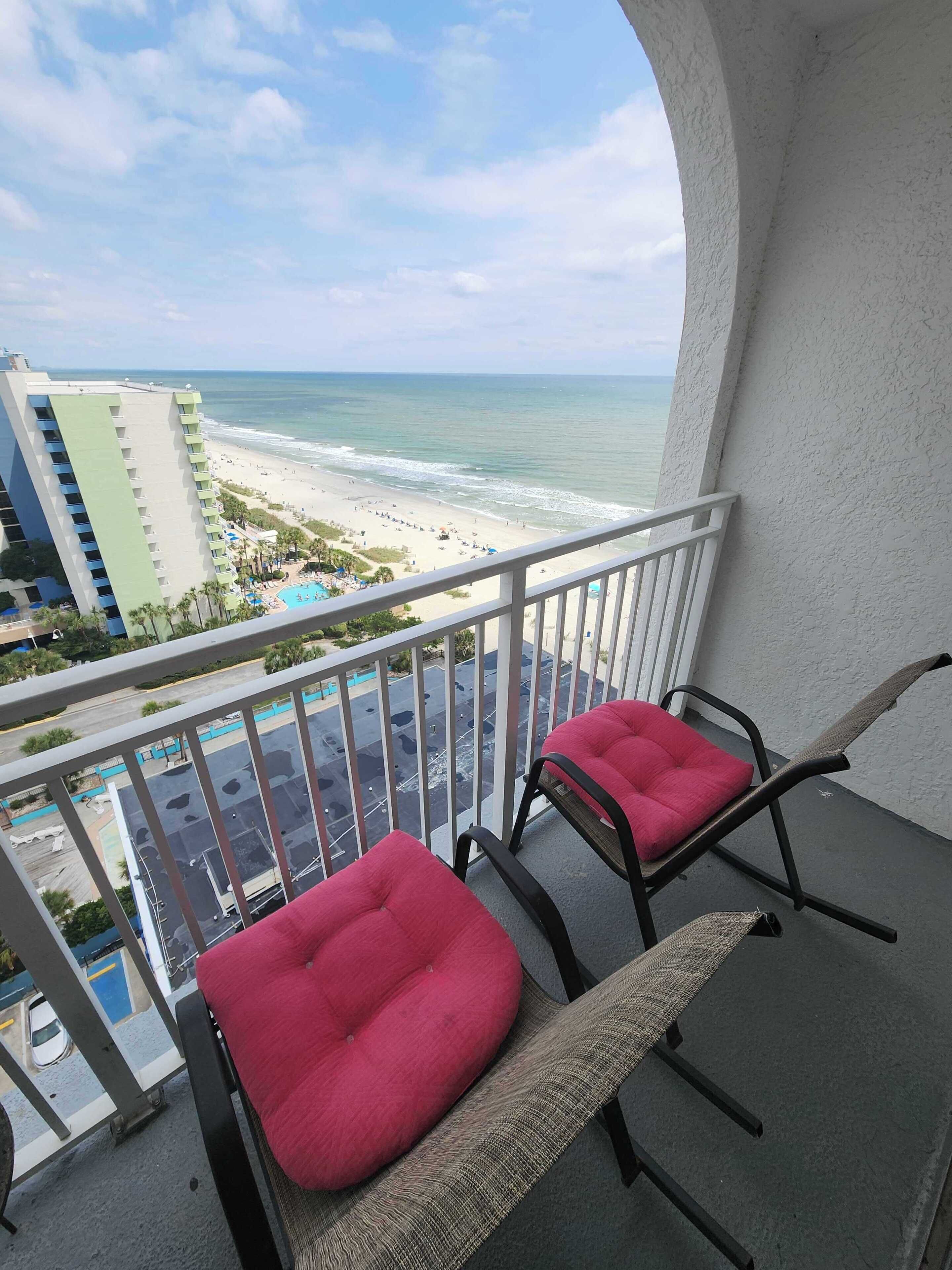 A balcony with two chairs, a view of the beach, and a parking lot.