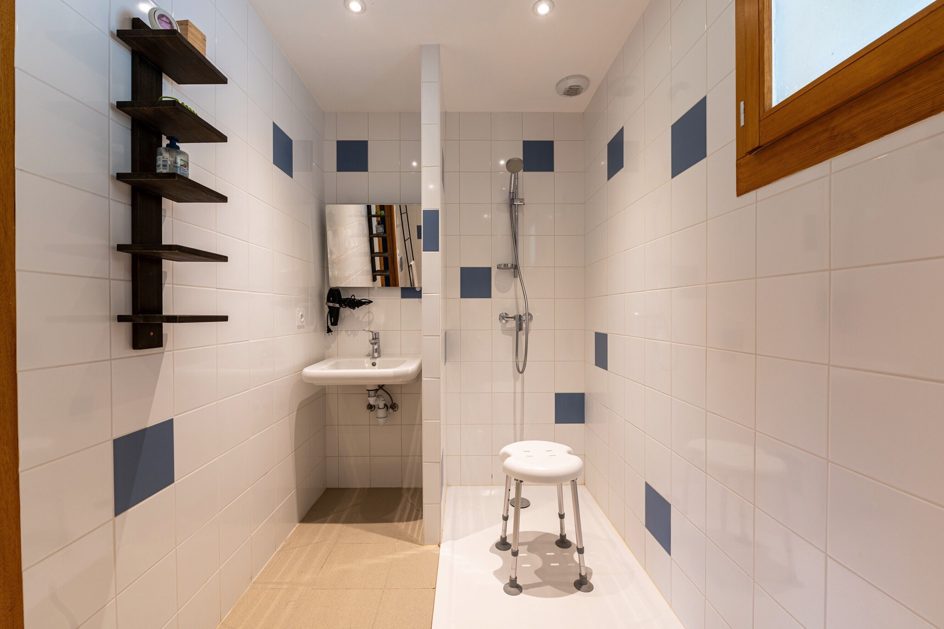 Un baño con lavamanos blanco, ducha y un taburete. Hay estantes fijos en la pared.