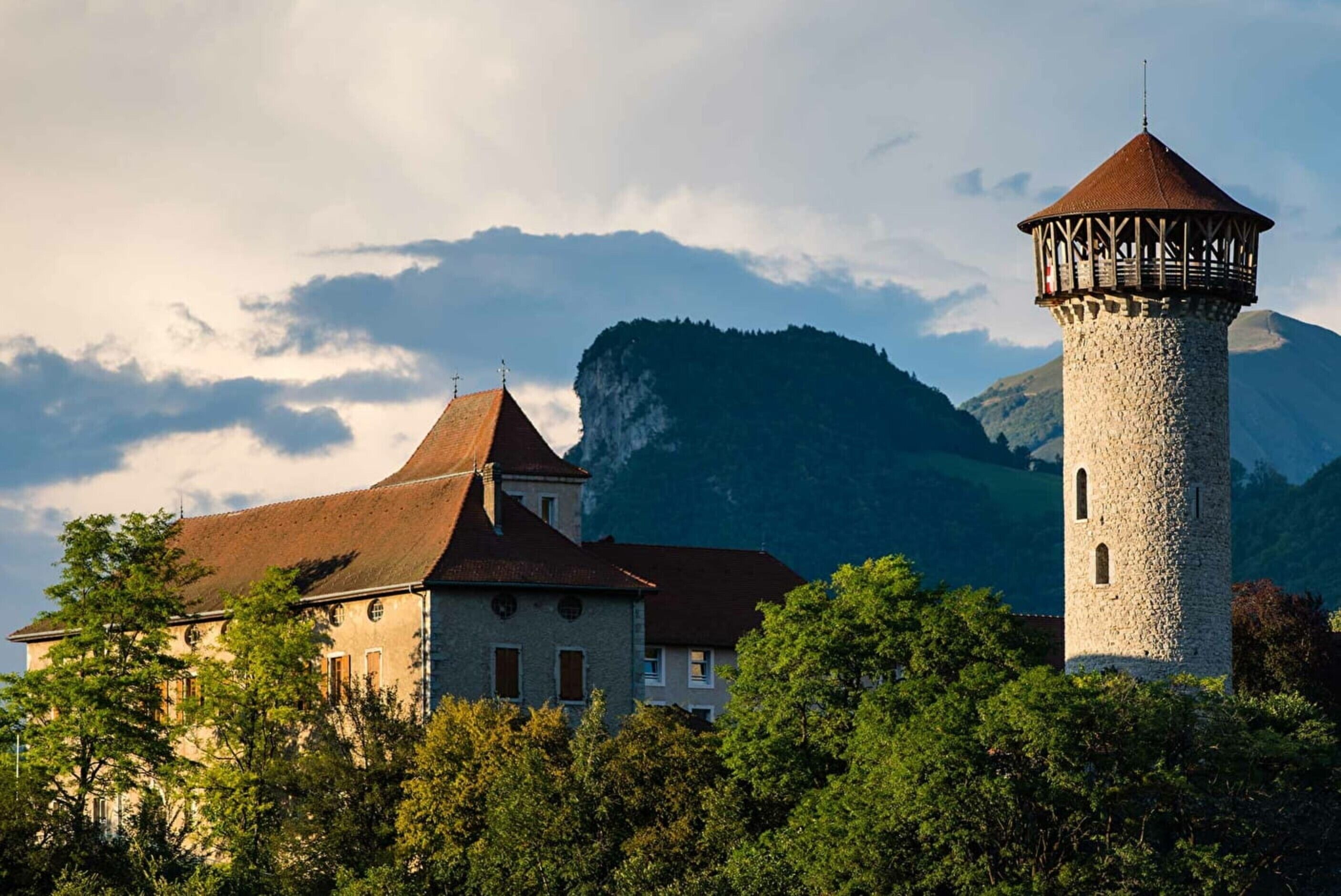 Ein steinerner Turm mit einem spitzen Dach, ein Gebäude mit roten Ziegeldächern und eine gebirgige Kulisse.