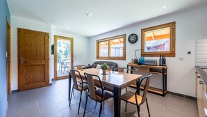 Een eetgedeelte met een houten tafel en stoelen, een klok aan de muur, en uitzicht op een gebouw door een raam.