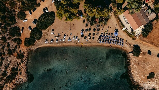 Ein Luftbild zeigt einen Strand mit einem großen, klaren Wasserkörper, einem Parkplatz mit Autos und einem Gebäude mit rotem Dach.