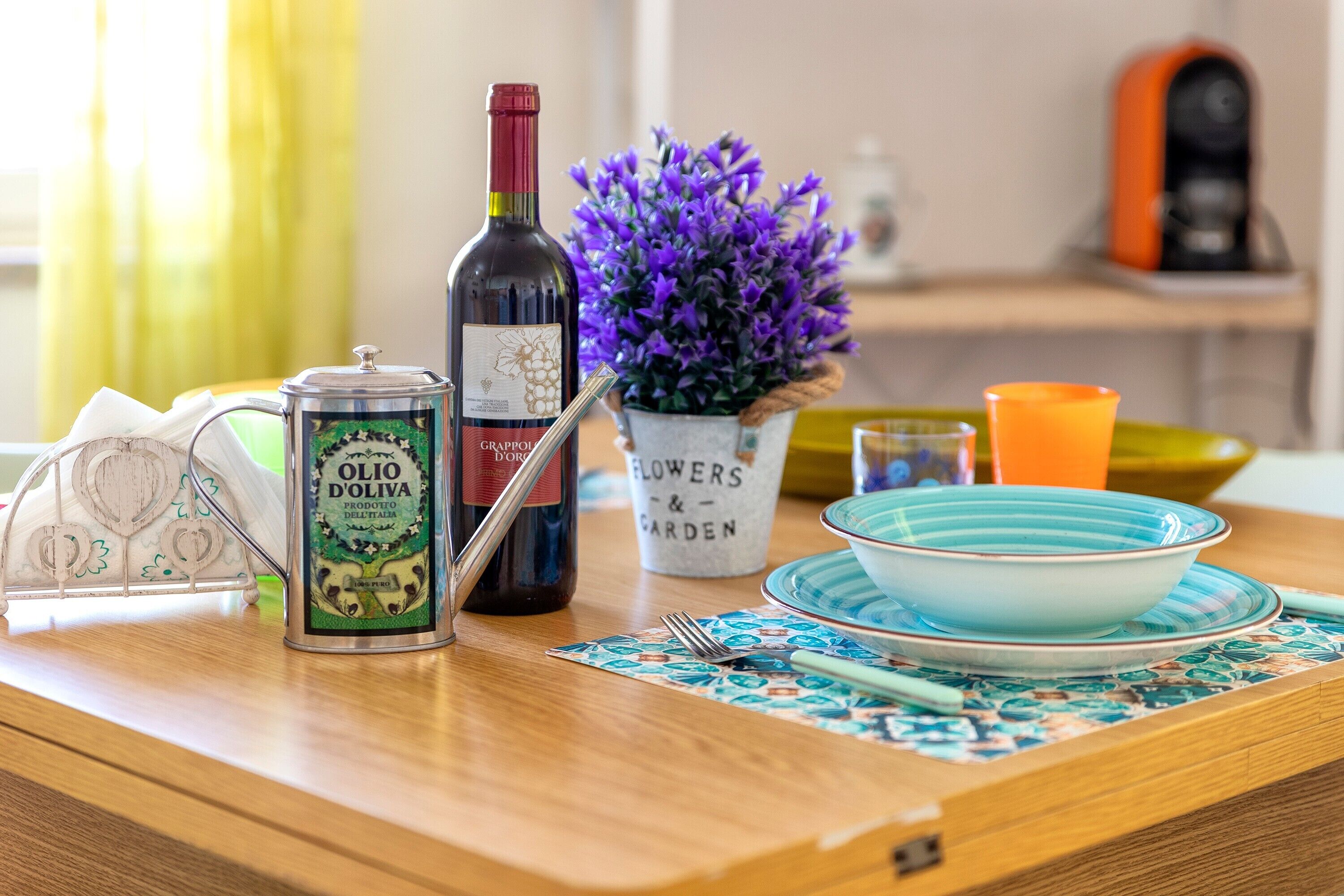 A kitchen counter with a wine bottle, a can of Olio D'Oliva, a potted plant, and a blue bowl on a patterned placemat.
