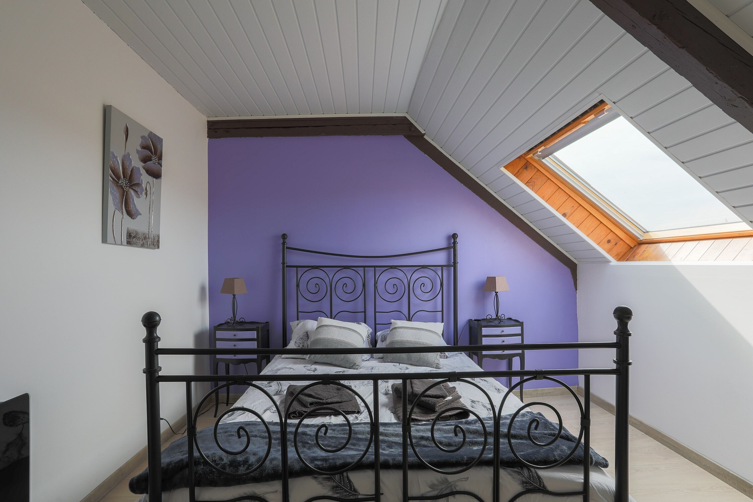 A bedroom with a sloped ceiling, a skylight, a bed with a metal frame, two bedside tables, and a purple wall.