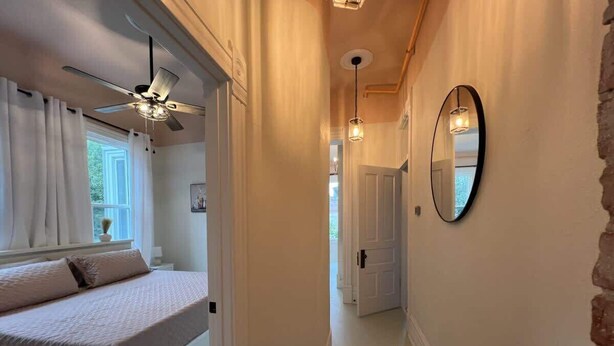 A bedroom with a bed, a ceiling fan, a window with curtains, and a hallway leading to another room.