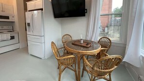 A compact kitchen with a white refrigerator, microwave, and oven, a round dining table with wicker chairs, and a flat-screen TV mounted on the wall.