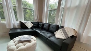 A modern living room with a black sectional sofa, a cream-colored ottoman, and sheer white curtains.