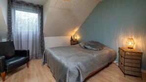 A bedroom with a bed, a chair, a nightstand with a lamp, and a window with curtains. - Holiday Home 'Upton 2 - Arradon Golfe Du Morbihan' with Private Terrace, Garden & Wi-Fi (Arradon)