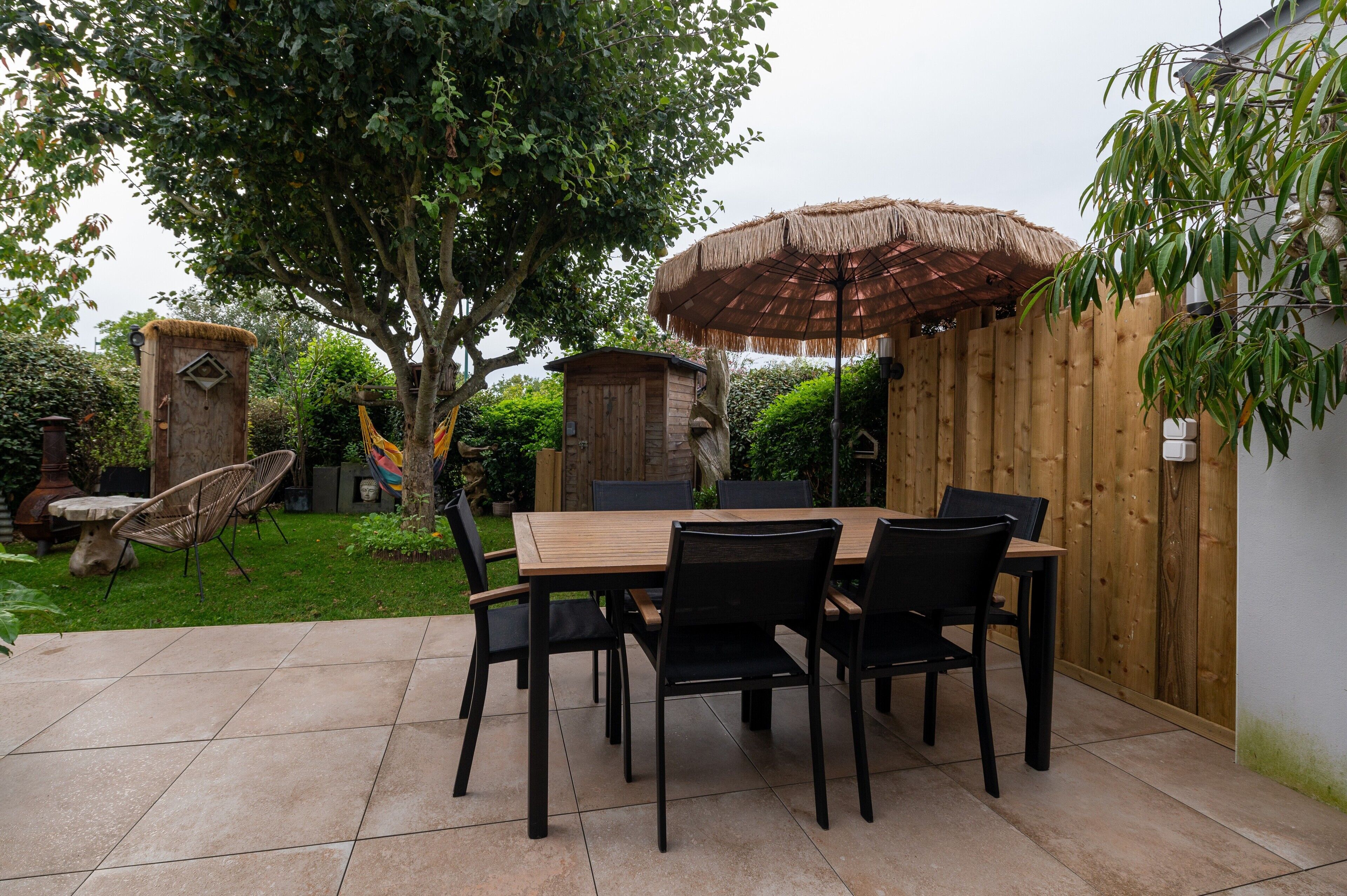 Een terras met een houten tafel en stoelen, een rieten zonnescherm en een houten omheining.