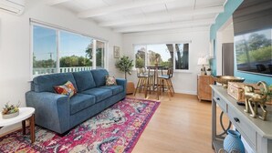 A spacious living area with a blue sofa, a colorful rug, a dining table with chairs, and a TV on the wall.