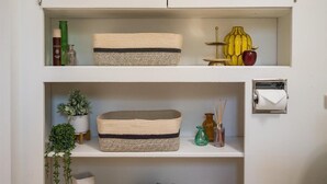 A white cabinet with two shelves, each containing woven baskets, a potted plant, and a roll of toilet paper.