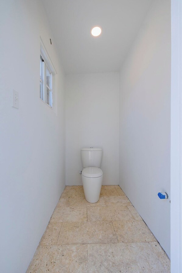 A small, narrow bathroom with a toilet, a window, and a tiled floor.