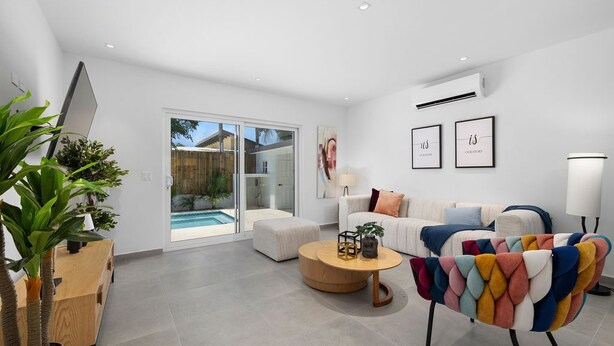 A modern living room with a sofa, a colorful armchair, a coffee table, and a TV. There is a sliding glass door leading to a pool area.