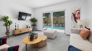 A modern living room with a flat-screen TV, a wooden coffee table, a sofa, and a large sliding glass door leading to a pool.