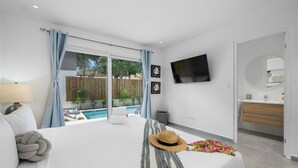 A modern bedroom with a large bed, a TV mounted on the wall, a view of a swimming pool, and a bathroom with a mirror and sink.