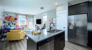 A modern kitchen with stainless steel appliances, a central island with a sink, and a dining area with a colorful sofa and a flat-screen TV.