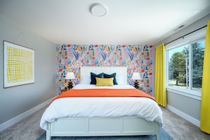 A bedroom with a bed, two bedside lamps, a window with yellow curtains, and a colorful floral wallpaper.