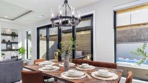 A modern dining area with a wooden table set for six, surrounded by brown chairs. A chandelier hangs above, and a large window offers a view of a hot tub. - Desert Sands at Pecan Valley Resort  (Hurricane)