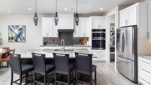 A modern kitchen with white cabinets, a central island with bar stools, stainless steel appliances, and pendant lighting. - Desert Sands at Pecan Valley Resort  (Hurricane)